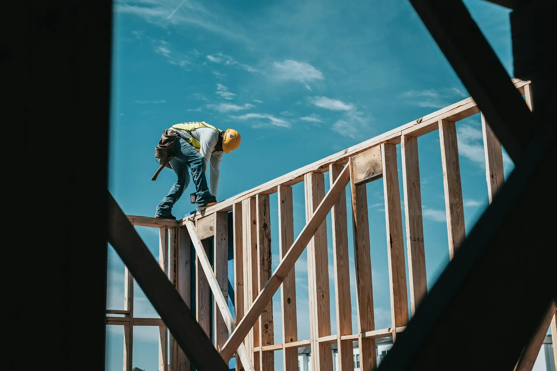A construction worker in a yellow hard hat and safety vest stands on the wooden frame of a building under construction against a blue sky.