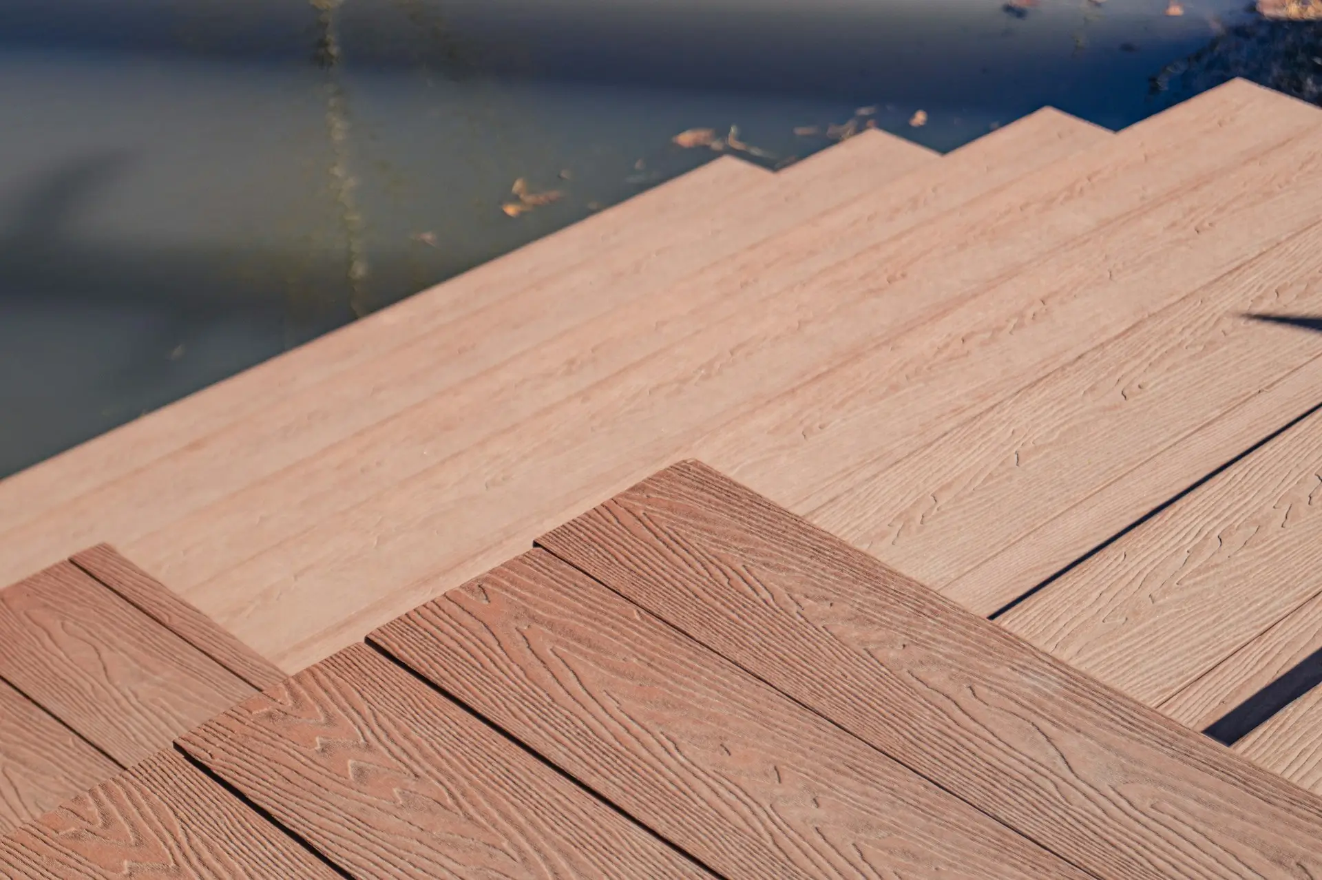 Wooden steps leading down to a body of water, with sunlight casting shadows and some leaves floating on the water’s surface.